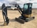 A compact excavator with a black and yellow body is parked on a dirt lot. The machinery has a large arm extending from its body, equipped with a digging bucket. The cabin is enclosed with large windows, and the tracks are visible beneath. In the background, there are other industrial vehicles and equipment visible, along with some structures and a cloudy sky.