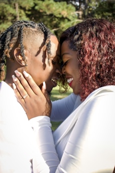 A couple embraces closely, pressing their foreheads together while smiling. They are bathed in natural light amidst a backdrop of green foliage. Both wear white attire and exhibit affectionate expressions. One person has intricate braided hair, and the other has curly hair with a ring visible on their hand.