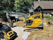 Two large excavators are engaged in a construction project near a house in a wooded area. The yellow excavator is prominent in the foreground, while the darker excavator is slightly behind it. They are surrounded by gravel and earth, suggesting site preparation or landscaping work.