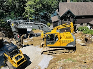 Two large excavators are engaged in a construction project near a house in a wooded area. The yellow excavator is prominent in the foreground, while the darker excavator is slightly behind it. They are surrounded by gravel and earth, suggesting site preparation or landscaping work.