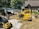 Two large excavators are engaged in a construction project near a house in a wooded area. The yellow excavator is prominent in the foreground, while the darker excavator is slightly behind it. They are surrounded by gravel and earth, suggesting site preparation or landscaping work.