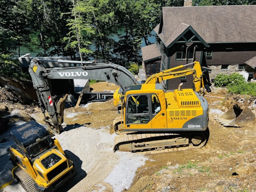 Two large excavators are engaged in a construction project near a house in a wooded area. The yellow excavator is prominent in the foreground, while the darker excavator is slightly behind it. They are surrounded by gravel and earth, suggesting site preparation or landscaping work.