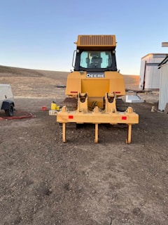 A large yellow construction vehicle, possibly a bulldozer, is positioned on a dirt area. The vehicle has a clearly visible grille and windows, with the brand name displayed prominently. There are additional structures nearby, including a trailer and some equipment.