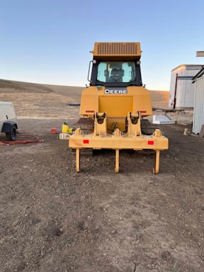 A large yellow construction vehicle, possibly a bulldozer, is positioned on a dirt area. The vehicle has a clearly visible grille and windows, with the brand name displayed prominently. There are additional structures nearby, including a trailer and some equipment.
