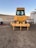 A large yellow construction vehicle, possibly a bulldozer, is positioned on a dirt area. The vehicle has a clearly visible grille and windows, with the brand name displayed prominently. There are additional structures nearby, including a trailer and some equipment.