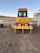 A large yellow construction vehicle, possibly a bulldozer, is positioned on a dirt area. The vehicle has a clearly visible grille and windows, with the brand name displayed prominently. There are additional structures nearby, including a trailer and some equipment.