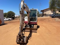 A construction site with a small excavator in the foreground, positioned on a dirt ground. In the background, there are trees, a white building with steps, and several parked vehicles, including a white truck and a silver car. The sky is clear and blue, and the surroundings have a dry and sunny atmosphere.