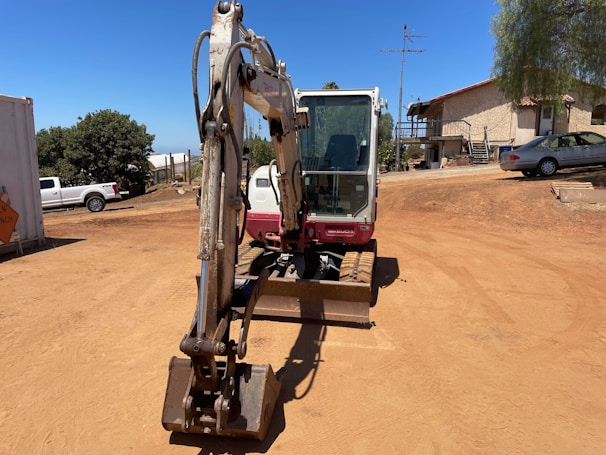 A construction site with a small excavator in the foreground, positioned on a dirt ground. In the background, there are trees, a white building with steps, and several parked vehicles, including a white truck and a silver car. The sky is clear and blue, and the surroundings have a dry and sunny atmosphere.