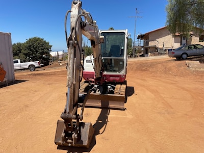 A construction site with a small excavator in the foreground, positioned on a dirt ground. In the background, there are trees, a white building with steps, and several parked vehicles, including a white truck and a silver car. The sky is clear and blue, and the surroundings have a dry and sunny atmosphere.