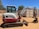 A compact excavator with a red and white color scheme is parked on a dirt surface in an outdoor setting. It has a cab with windows, a metal arm, and a digging bucket. In the background, there are trees and large shipping containers.