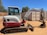 A compact excavator with a red and white color scheme is parked on a dirt surface in an outdoor setting. It has a cab with windows, a metal arm, and a digging bucket. In the background, there are trees and large shipping containers.