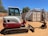 A compact excavator with a red and white color scheme is parked on a dirt surface in an outdoor setting. It has a cab with windows, a metal arm, and a digging bucket. In the background, there are trees and large shipping containers.