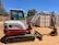A compact excavator with a red and white color scheme is parked on a dirt surface in an outdoor setting. It has a cab with windows, a metal arm, and a digging bucket. In the background, there are trees and large shipping containers.