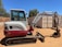 A compact excavator with a red and white color scheme is parked on a dirt surface in an outdoor setting. It has a cab with windows, a metal arm, and a digging bucket. In the background, there are trees and large shipping containers.