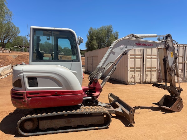 A compact excavator with a red and white color scheme is parked on a dirt surface in an outdoor setting. It has a cab with windows, a metal arm, and a digging bucket. In the background, there are trees and large shipping containers.
