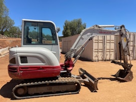 A compact excavator with a red and white color scheme is parked on a dirt surface in an outdoor setting. It has a cab with windows, a metal arm, and a digging bucket. In the background, there are trees and large shipping containers.