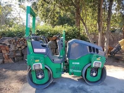 A green double drum compactor is parked on a dirt surface next to a stack of cut logs and surrounded by trees. The vehicle has visible identification numbers and logos on its side, and the surrounding area has scattered sunlight filtering through the trees.