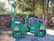 A green double drum compactor is parked on a dirt surface next to a stack of cut logs and surrounded by trees. The vehicle has visible identification numbers and logos on its side, and the surrounding area has scattered sunlight filtering through the trees.
