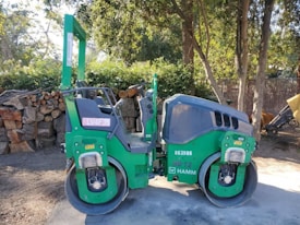 A green double drum compactor is parked on a dirt surface next to a stack of cut logs and surrounded by trees. The vehicle has visible identification numbers and logos on its side, and the surrounding area has scattered sunlight filtering through the trees.