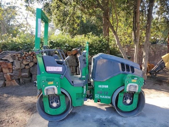 A green double drum compactor is parked on a dirt surface next to a stack of cut logs and surrounded by trees. The vehicle has visible identification numbers and logos on its side, and the surrounding area has scattered sunlight filtering through the trees.