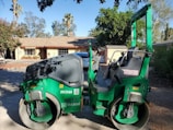 A green asphalt roller machine is parked on a driveway in front of a suburban house with a light-colored exterior. The house has a sloped shingle roof and a visible chimney. The surroundings include trees and shrubs, contributing to a calm neighborhood setting.