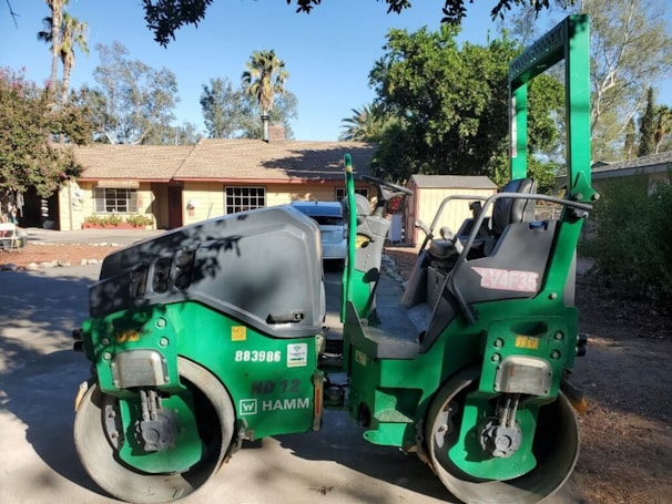 A green asphalt roller machine is parked on a driveway in front of a suburban house with a light-colored exterior. The house has a sloped shingle roof and a visible chimney. The surroundings include trees and shrubs, contributing to a calm neighborhood setting.