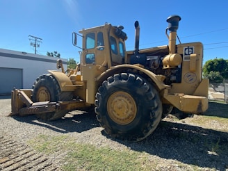 A large industrial tractor with thick wheels parked on gravel, bathed in sunlight with a shadow visible on the ground. The tractor is yellow and has a metal exhaust pipe protruding from its upper section. Nearby, a building is visible along with some power lines and trees in the background.