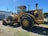 A large industrial tractor with thick wheels parked on gravel, bathed in sunlight with a shadow visible on the ground. The tractor is yellow and has a metal exhaust pipe protruding from its upper section. Nearby, a building is visible along with some power lines and trees in the background.