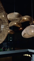A collection of woven rattan light fixtures hangs from the ceiling, casting warm light in the room. The setting appears to be a modern indoor space with dark walls and large windows.