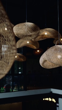 A collection of woven rattan light fixtures hangs from the ceiling, casting warm light in the room. The setting appears to be a modern indoor space with dark walls and large windows.