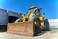 A large yellow bulldozer is parked on a gravel surface near a fenced industrial area. Its massive front blade is rusty, and large tires are prominent. An open shipping container and another vehicle can be seen in the background under a clear blue sky.
