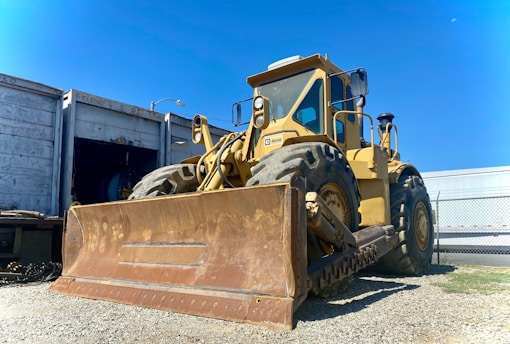 A large yellow bulldozer is parked on a gravel surface near a fenced industrial area. Its massive front blade is rusty, and large tires are prominent. An open shipping container and another vehicle can be seen in the background under a clear blue sky.