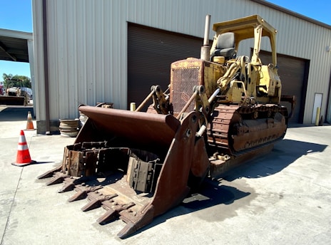 A large, yellow bulldozer with a rusted metal front scoop is parked on a concrete surface in front of a metal industrial building. Several orange and white traffic cones are placed nearby, and the area is well-lit with clear shadows indicating bright sunlight.