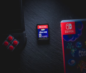A Nintendo Switch game cartridge labeled 'Cosmic Shake' is positioned at the center, surrounded by partial views of a Joy-Con controller on the left and a game case on the right. The scene is set against a dark background, highlighting the vibrant colors of the gaming elements.