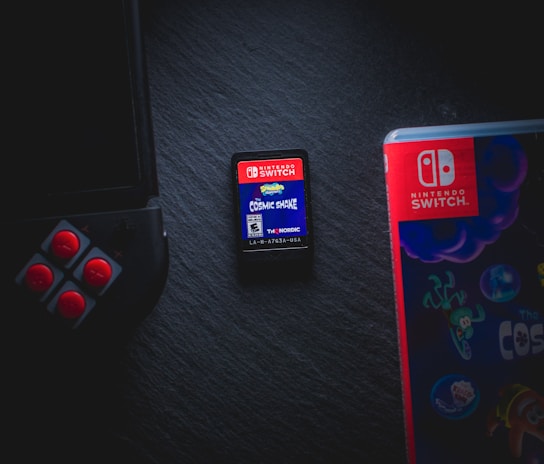 A Nintendo Switch game cartridge labeled 'Cosmic Shake' is positioned at the center, surrounded by partial views of a Joy-Con controller on the left and a game case on the right. The scene is set against a dark background, highlighting the vibrant colors of the gaming elements.