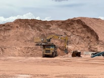 A large construction site featuring an excavator loading soil into a dump truck, with a background of sandy, excavated terrain. The scene suggests ongoing earthmoving or mining activity under a cloudy sky.