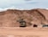 A large construction site featuring an excavator loading soil into a dump truck, with a background of sandy, excavated terrain. The scene suggests ongoing earthmoving or mining activity under a cloudy sky.