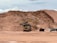 A large construction site featuring an excavator loading soil into a dump truck, with a background of sandy, excavated terrain. The scene suggests ongoing earthmoving or mining activity under a cloudy sky.