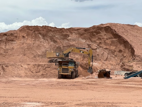 A large construction site featuring an excavator loading soil into a dump truck, with a background of sandy, excavated terrain. The scene suggests ongoing earthmoving or mining activity under a cloudy sky.