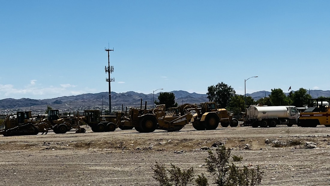 A lineup of large construction vehicles and machinery is positioned on a dirt lot under a clear blue sky. In the background, a series of mountains can be seen, along with some trees and a cell tower. The scene has a somewhat industrial and rugged atmosphere.