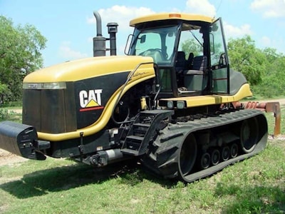 A Caterpillar agricultural tractor with large rubber tracks is parked on grass. The machine has a sleek yellow and black design, with the recognizable CAT logo on the side. The tractor features a spacious cabin with large windows and various machinery parts are visible.