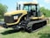 A Caterpillar agricultural tractor with large rubber tracks is parked on grass. The machine has a sleek yellow and black design, with the recognizable CAT logo on the side. The tractor features a spacious cabin with large windows and various machinery parts are visible.