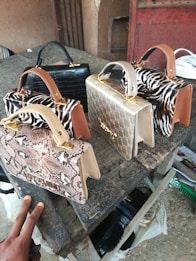 A collection of four stylish handbags placed on a rough wooden surface. They display a range of textures and colors, including snake print, zebra pattern, gold with embossed details, and black faux croc. The patterns and textures give off a luxurious and fashionable vibe.