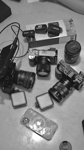 A collection of camera equipment and accessories including several cameras with attached lenses, a couple of additional lenses, wireless microphone systems, two square lights, and a smartphone with a transparent case. The items are arranged on a light-colored surface.