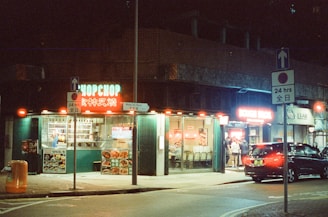 A lively nighttime street scene depicts a small restaurant with bright neon signs and illuminated windows. Several people are seen inside, and there’s a car parked nearby under the streetlights. The signage includes English and Chinese characters, contributing to an urban, multicultural atmosphere.