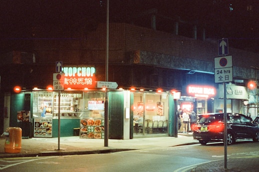 A lively nighttime street scene depicts a small restaurant with bright neon signs and illuminated windows. Several people are seen inside, and there’s a car parked nearby under the streetlights. The signage includes English and Chinese characters, contributing to an urban, multicultural atmosphere.