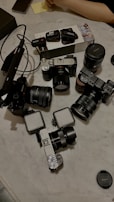 A collection of camera equipment is arranged on a marble surface. The setup includes multiple cameras, lenses, microphone packs, and a camera lens cap. A person's arm is partially visible in the upper right corner, near a piece of paper.