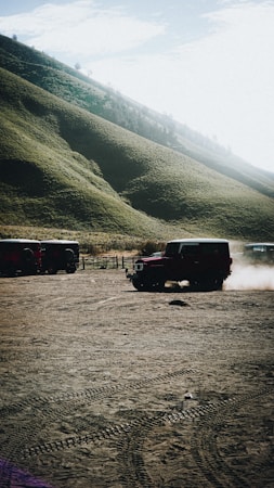 A rugged, muddy landscape with a steep, grassy hill in the background. A red off-road vehicle is driving across the dirt field, creating clouds of dust. Other similar vehicles are parked nearby under a bright, slightly cloudy sky.