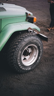 A close-up view of the front left tire and fender of a light green off-road vehicle, possibly a Jeep, parked on a sandy surface. The tire is a rugged Bridgestone Dueler, suitable for off-road conditions. The background shows part of a person wearing dark pants and tan boots.