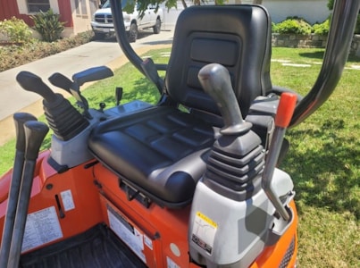 A close-up view of the control seat area of a small construction vehicle, likely an excavator or similar heavy machinery. The image prominently features a black padded seat with multiple levers and controls around it. The background shows a grassy area, a paved walkway, and a parked white van near a house.